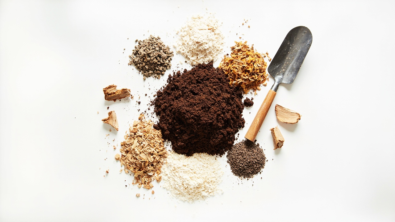 A flat-lay of monstera soil ingredients including a central pile of dark potting mix surrounded by smaller piles of perlite, orchid bark, coco coir, worm castings, and pine bark pieces, with a small hand trowel on a white surface.